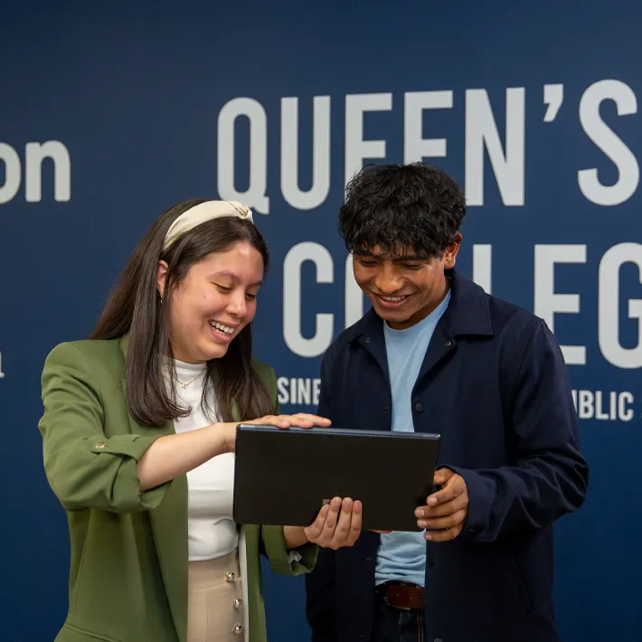 A photo of two students standing in front of campus mural, looking at iPad and smiling.