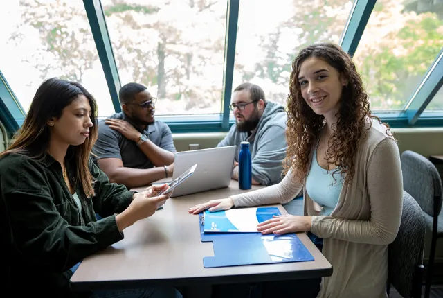 A group Students sitting at a table in the common area on campus using laptop, folder and ipad.