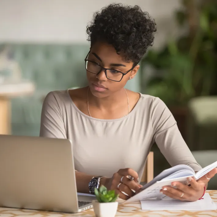 A woman sitting at desk researching on computer and looking through notes.