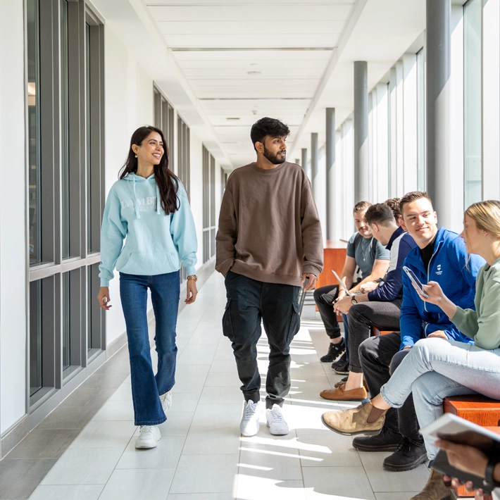 Two students walking down the Sarnia Campus hallway with students sitting on the benches.