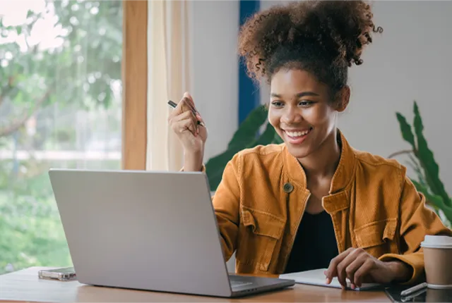 A women sitting in front of a laptop at desk with a window next to her and a coffee cup.