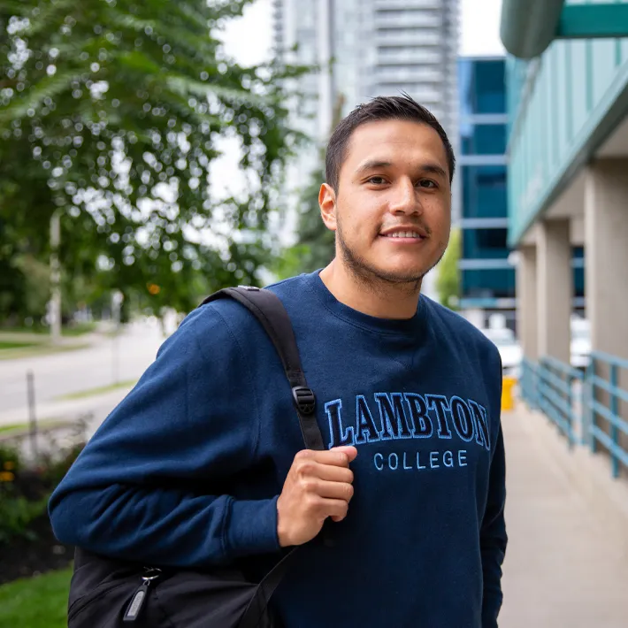 A photo of a student wearing a packback and blue sweater standing outside of campus.