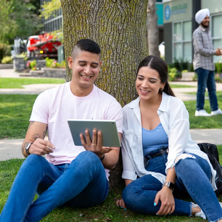 A lifestyle photo of two students wearing white tshirts and blue jeans sitting outside against a tree and looking at ipad together.