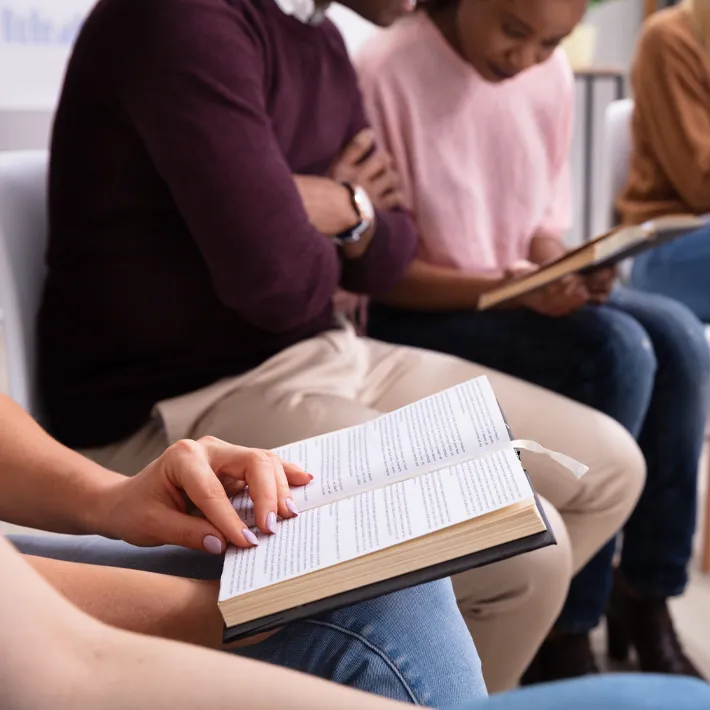 A close-up photo of a student holding a book with others sitting next her also reading a book.