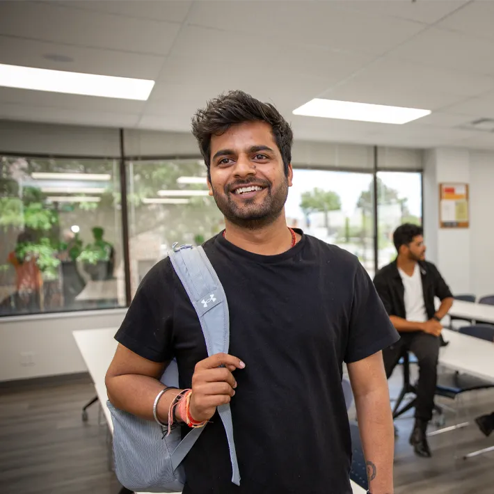 A student posing for a photo in a classroom, smiling and carrying backpack on shoulder.