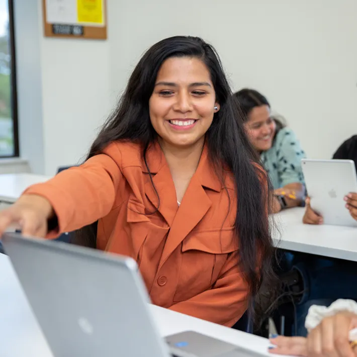 An international student in a classroom sitting at a desk pointing at computer screen.