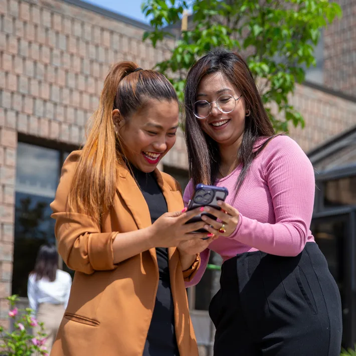 Two international students standing outside looking at a phone and smiling.