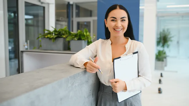 A student standing in hotel lobby holding a clipboard.