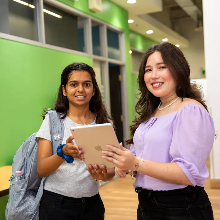 Two international students posing for photo on campus holding an ipad and smiling.