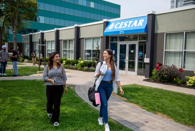 Two students walking away from the Cestar campus doors, talking, causal.