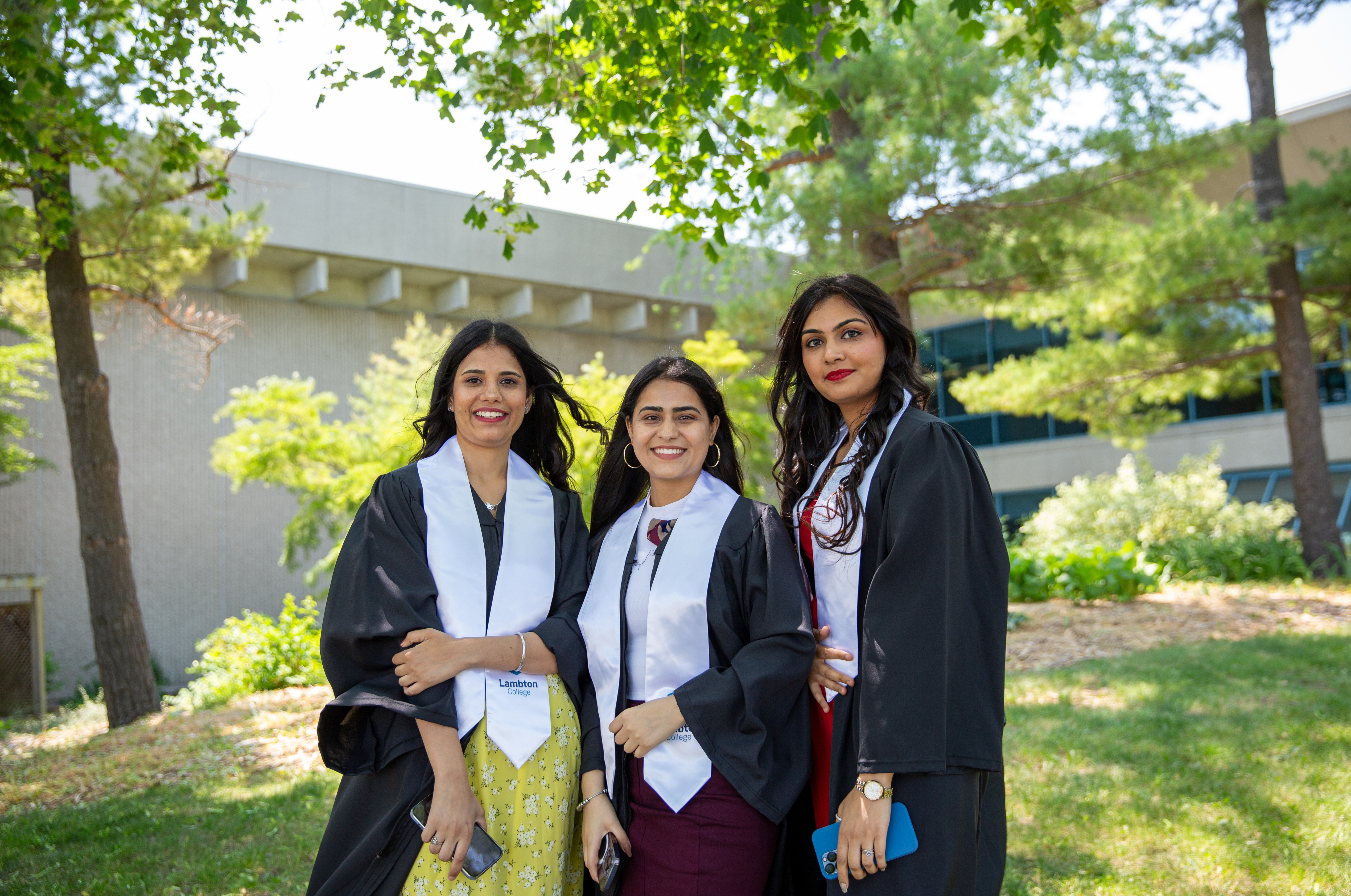 Three women international students posing for photo outside wearing convocation gowns.