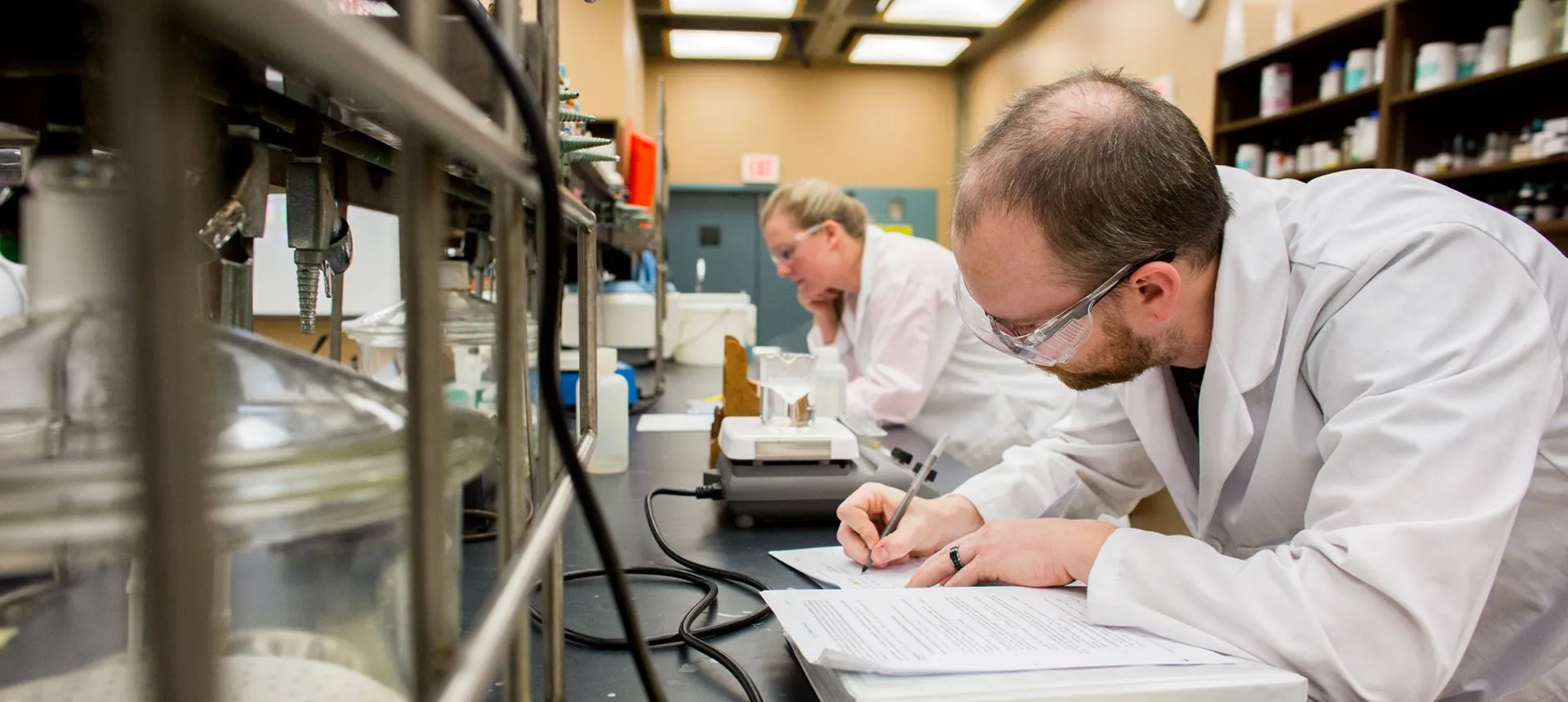 Student in lab coat taking notes at a lab desk